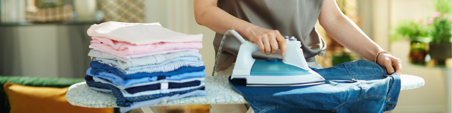 Een persoon strijkt een spijkerbroek op een strijkplank, naast een nette stapel schone en gestreken kleding. De omgeving is huiselijk en goed verlicht.